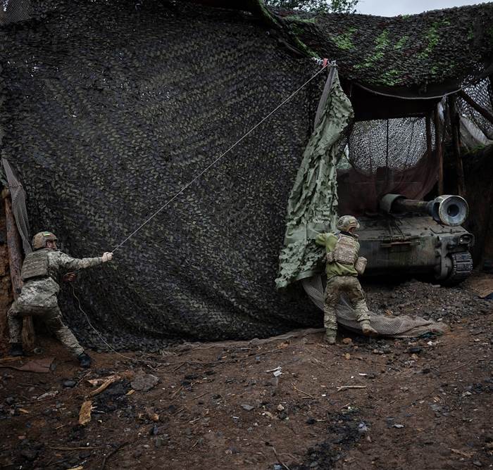 Solders in fatigues pulling back a camouflaged curtain hiding artillery in the field.