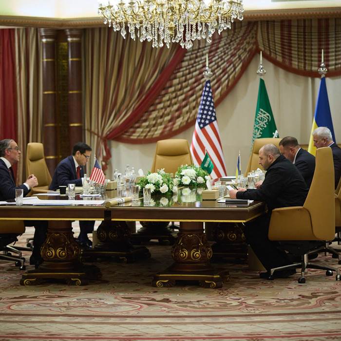 Men siting at a large table in a room with thick curtains and multiple chandeliers.