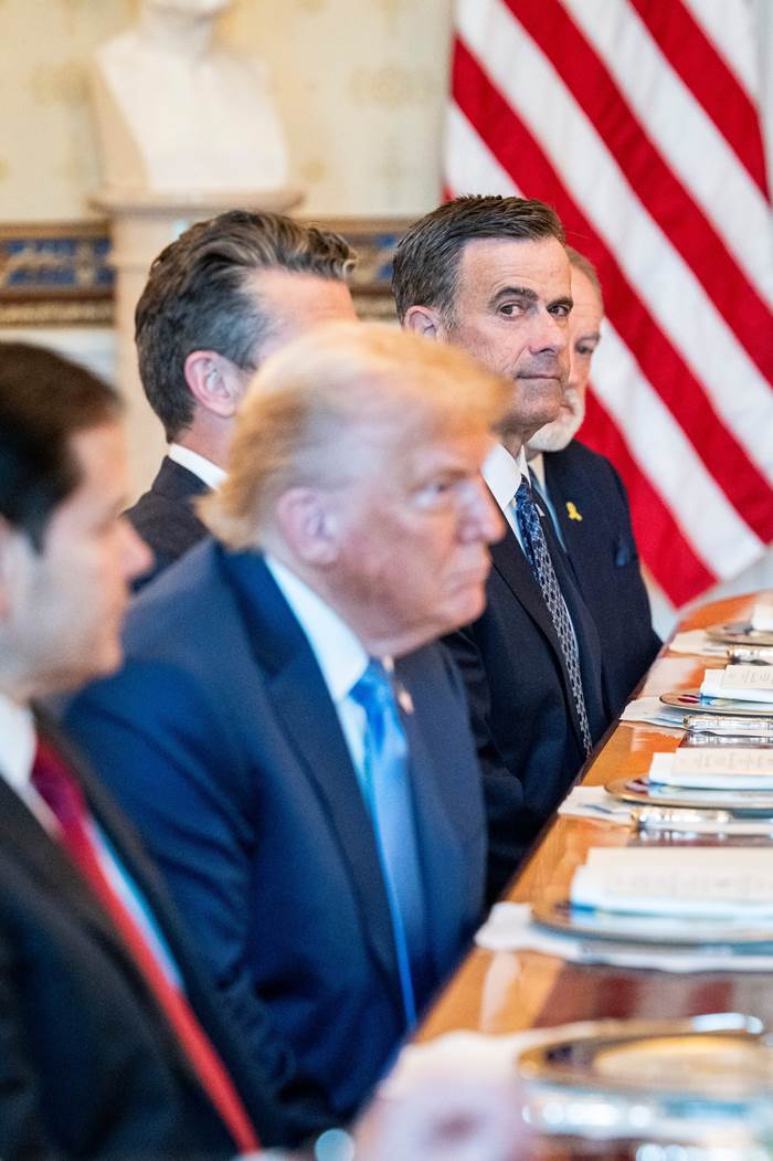 Suited men sitting at a table with the American flag in the background.